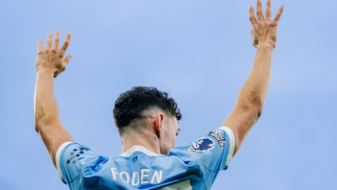 Phil Foden, wearing a Manchester City jersey, raises his arms in celebration. Visible details include the Premier League badge.