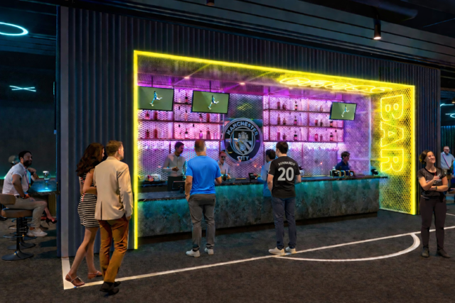 People standing at a modern bar with Manchester City logo, neon lights, and sports screens above.