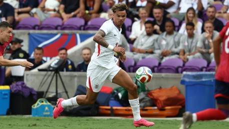A soccer player in white jersey running with the ball during a match, with spectators in the background.
