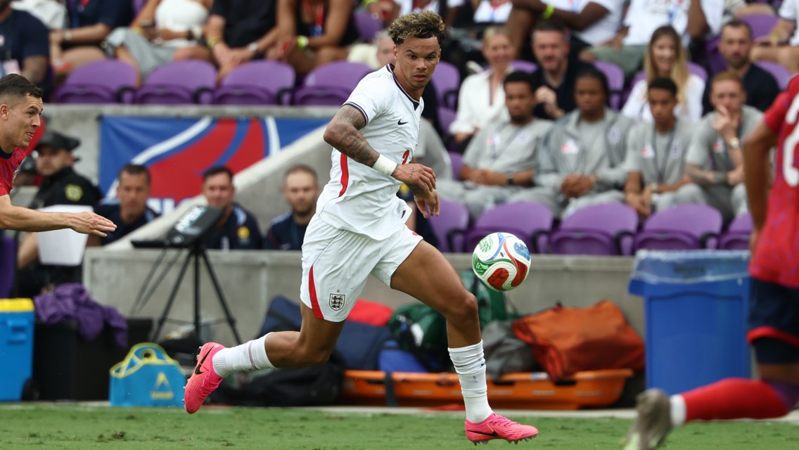 A soccer player in white jersey running with the ball during a match, with spectators in the background.