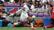 A soccer player in white jersey running with the ball during a match, with spectators in the background.