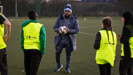 A coach holding a soccer ball instructs young players wearing Premier League training vests on an outdoor training field.