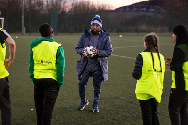 A coach holding a soccer ball instructs young players wearing Premier League training vests on an outdoor training field.