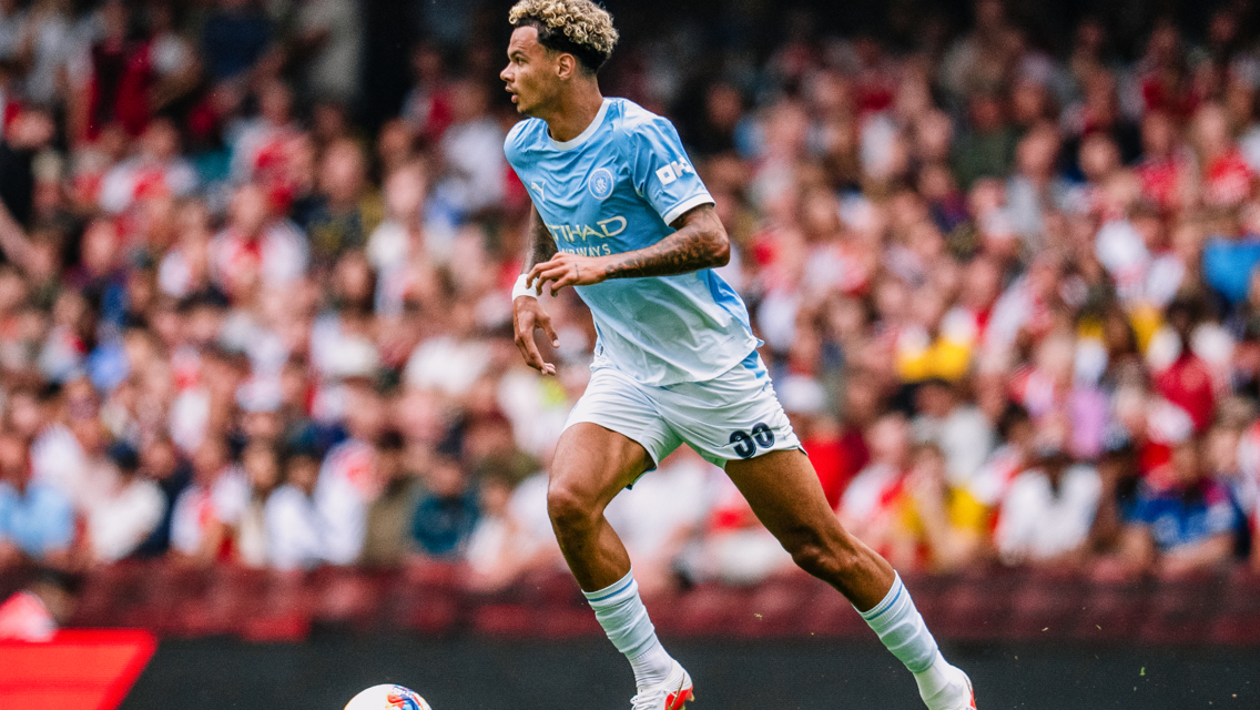 A soccer player in a Manchester City kit is running on the field during a match. The crowd is blurred in the background.
