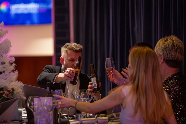 A blurred image of a person pouring beer at a Christmas-themed party, surrounded by others holding drinks, with a white Christmas tree and a Manchester Ship logo visible in the background.
