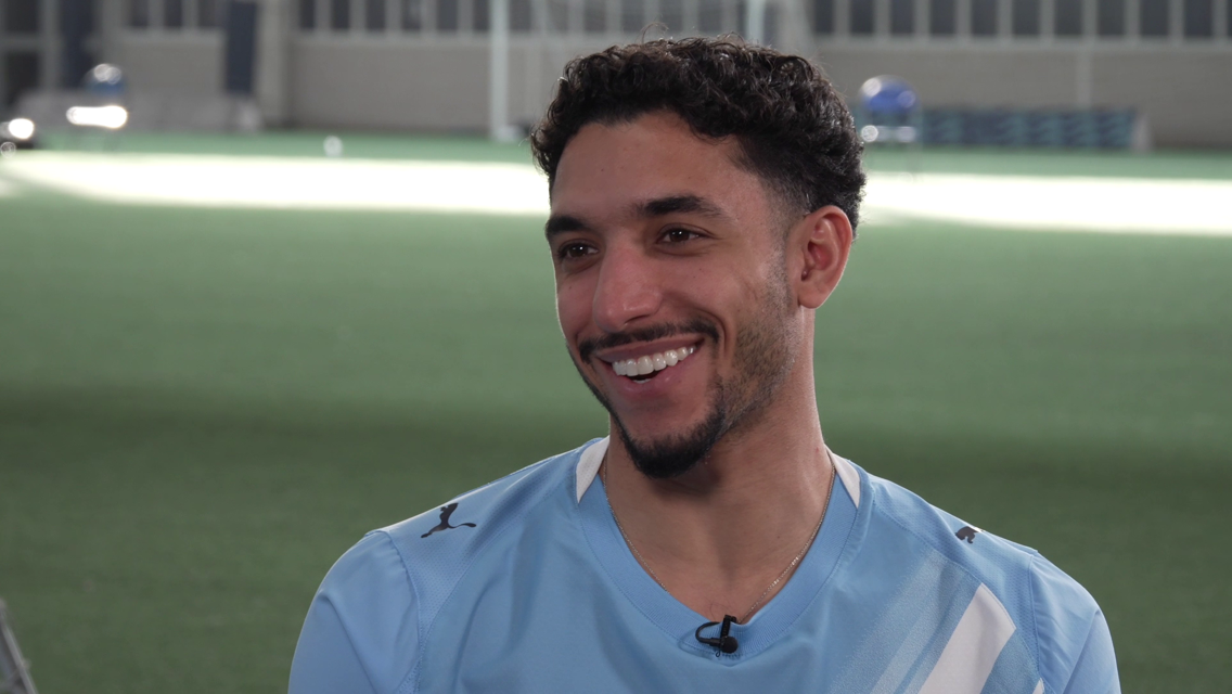 A person in a blurred photo wears a light blue Manchester City kit standing in an indoor pitch.