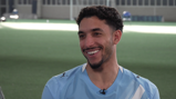 A person in a blurred photo wears a light blue Manchester City kit standing in an indoor pitch.