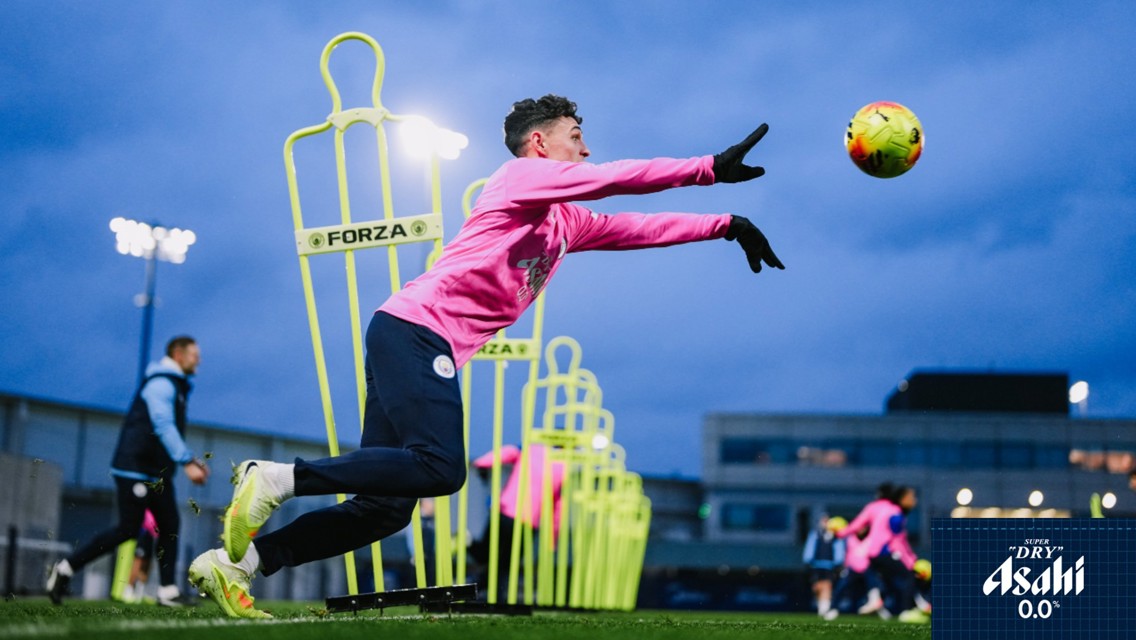 A football player in a pink Manchester City training kit practices diving to catch a ball, with training mannequins labeled 'FORZA' in the background, under an evening sky.