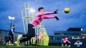 A football player in a pink Manchester City training kit practices diving to catch a ball, with training mannequins labeled 'FORZA' in the background, under an evening sky.