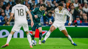 A football match moment showing a player from Manchester City in a dark teal kit dribbling against two Real Madrid players in white kits during the UEFA Champions League.