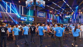 A lively fan event at City Hall with people wearing Manchester City jerseys. Large screens and colorful lighting create a vibrant atmosphere.