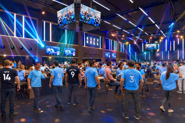 A lively fan event at City Hall with people wearing Manchester City jerseys. Large screens and colorful lighting create a vibrant atmosphere.