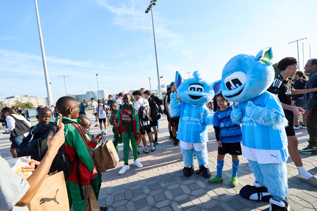 Manchester City mascots Moonchester and Moonbeam pose with children and fans at an outdoor event, likely related to the club's community activities.
