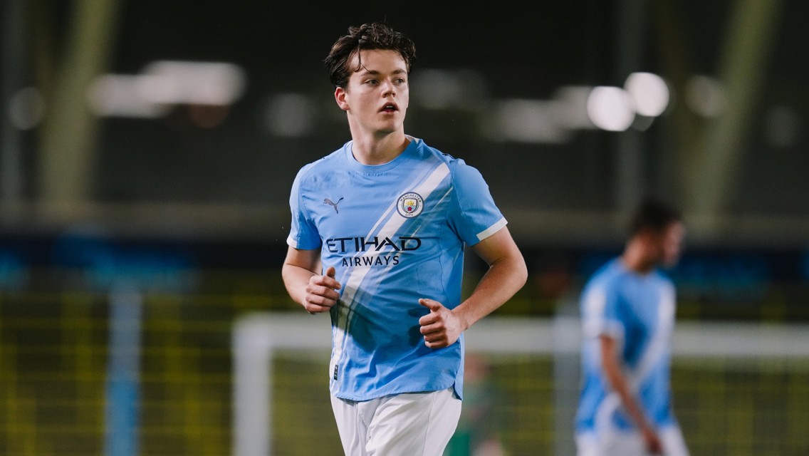 A football player wearing a Manchester City kit during a match, running on the field with blurred teammates in the background.