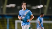 A football player wearing a Manchester City kit during a match, running on the field with blurred teammates in the background.