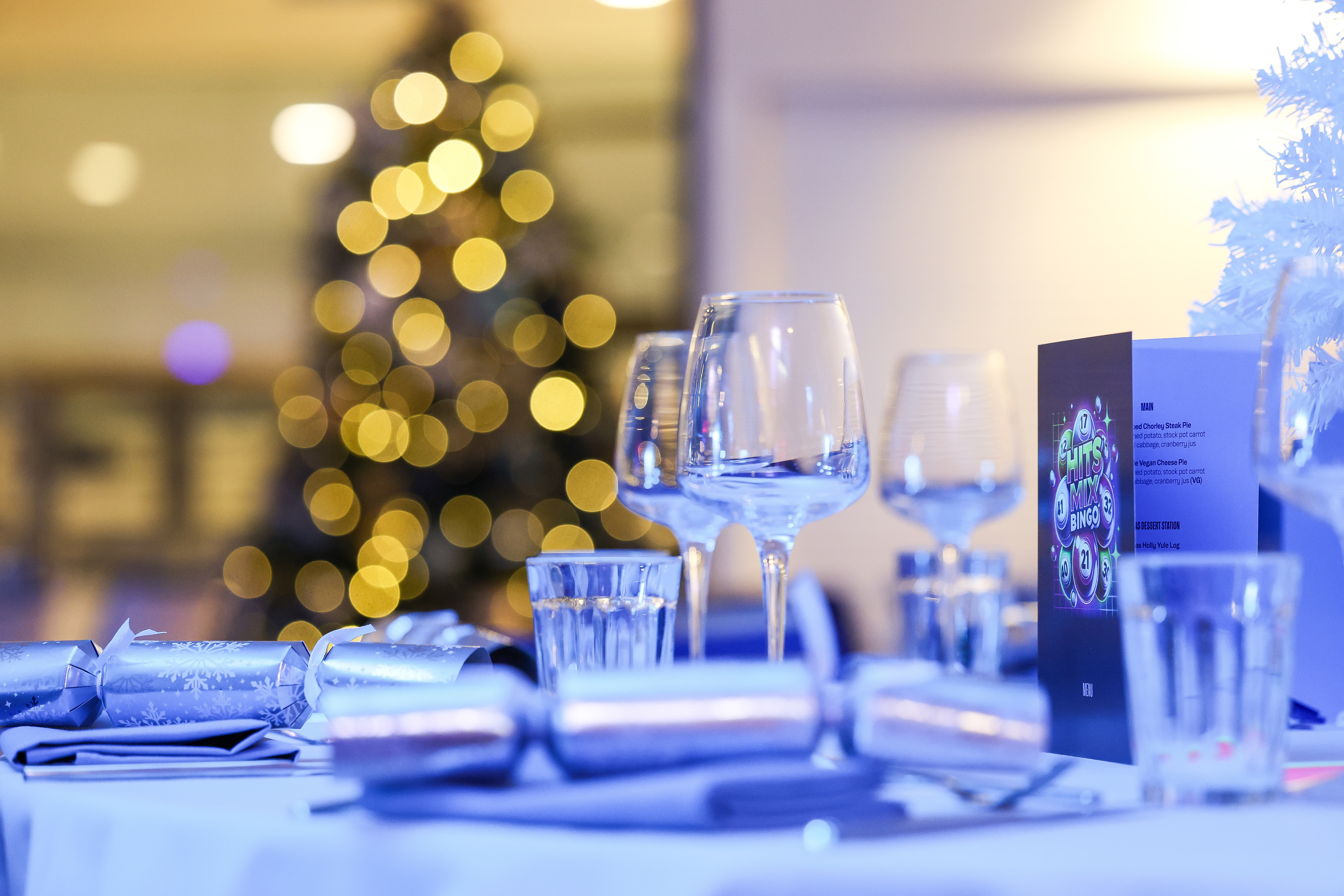 A dining table set with glassware and decorations, featuring a menu card and Christmas crackers. A Christmas tree with bokeh lights is visible in the background.