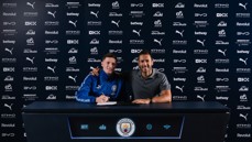 Two people at a table with a Manchester City logo backdrop. One person wearing a blue jacket appears to be signing a document.