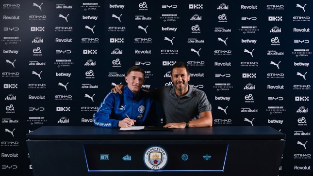 Two people at a table with a Manchester City logo backdrop. One person wearing a blue jacket appears to be signing a document.