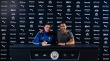 Two people at a table with a Manchester City logo backdrop. One person wearing a blue jacket appears to be signing a document.