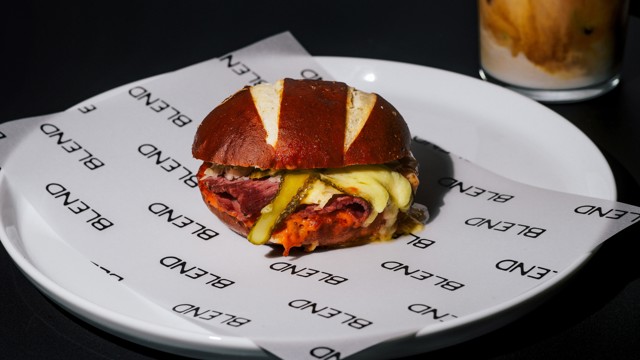 A pretzel bun burger with cheese, pickles, and pastrami on a branded paper-lined plate.