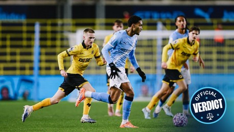 A football player in a light blue kit running with the ball pursued by players in yellow kits on a field. 'Official Verdict' badge on the bottom right.
