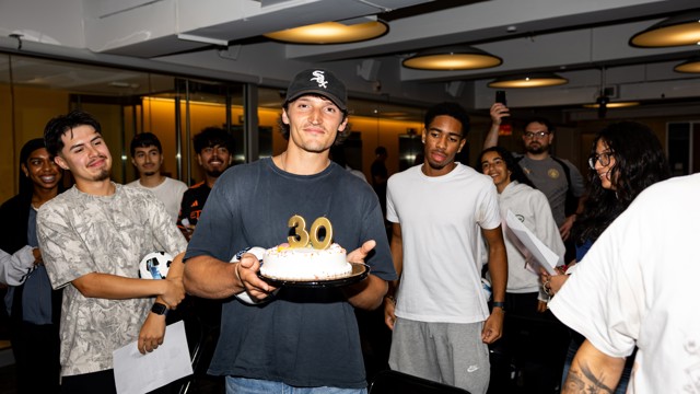 A group of people celebrate a birthday with a cake displaying the number 30 and smile. The person holding the cake is wearing a black cap and a dark shirt.