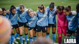 A group of Manchester City women players huddling on a soccer field wearing blue jerseys. Text says 'Thank You Laura.'