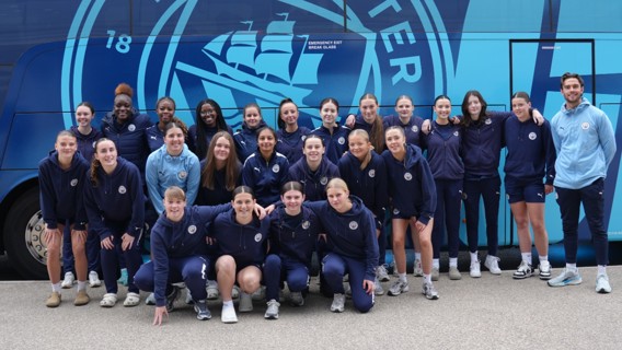 A group of people in Manchester City attire posing in front of a team bus with the club logo.