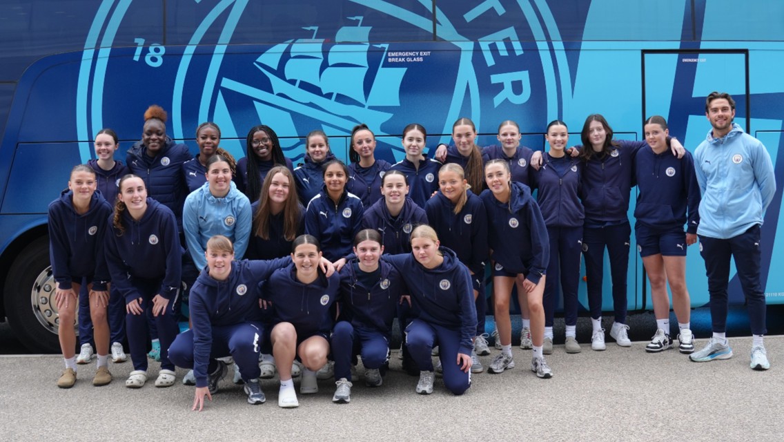 A group of people in Manchester City attire posing in front of a team bus with the club logo.