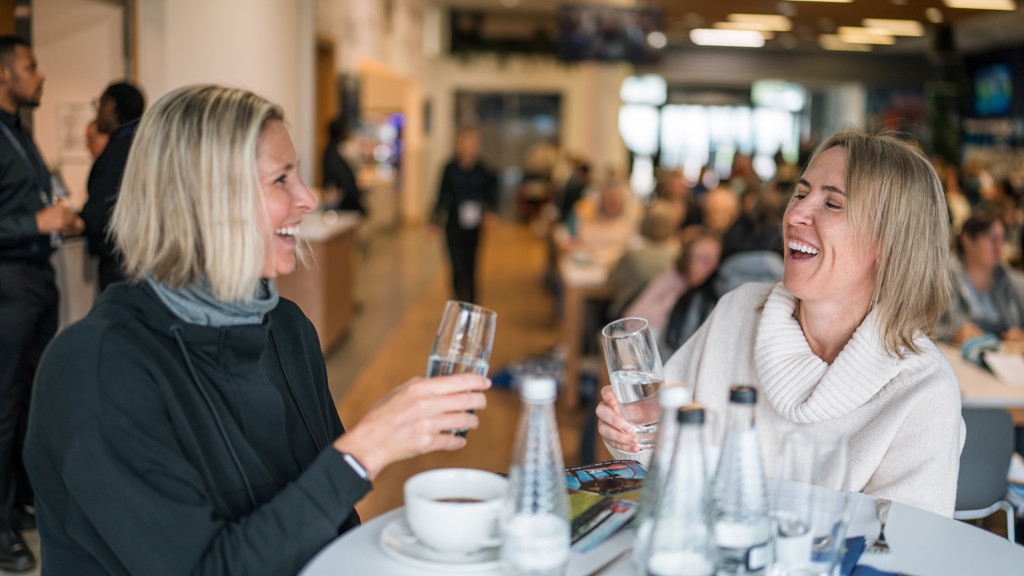 Two women with blurred faces sitting at a table in a busy lounge, holding glasses of water, with water bottles and a cup in front of them. The background shows other people in a hospitality setting.