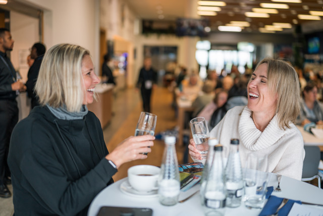 Two women with blurred faces sitting at a table in a busy lounge, holding glasses of water, with water bottles and a cup in front of them. The background shows other people in a hospitality setting.