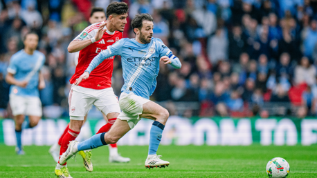 Football match action with players from Manchester City in blue and Arsenal in red during a game, with a crowd in the background.