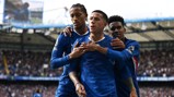 Three Chelsea players wearing blue jerseys celebrate a goal with arms wrapped around each other at a stadium filled with spectators.