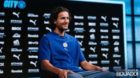 A person wearing a blue Manchester City shirt sits for an interview. The backdrop features sponsor logos like Puma, Etihad, and Revolut.