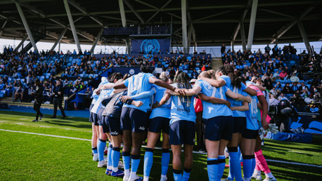 A group of women soccer players in blue jerseys huddling on the field with an audience in the background.