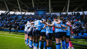 A group of women soccer players in blue jerseys huddling on the field with an audience in the background.