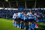 A group of women soccer players in blue jerseys huddling on the field with an audience in the background.