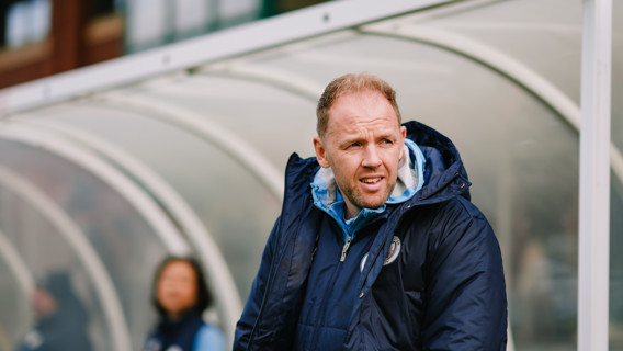 A man wearing a navy blue jacket and a light blue hoodie stands near a sideline, likely coaching. The jacket has a Manchester City logo.