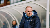A man wearing a navy blue jacket and a light blue hoodie stands near a sideline, likely coaching. The jacket has a Manchester City logo.