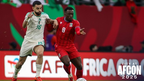 Two soccer players in action during an AFCON match, one wearing a white jersey and the other wearing a red jersey. AFCON 2025 branding is visible.