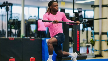 Athlete in a pink jersey and navy shorts performing a resistance exercise with a bar in a gym setting.