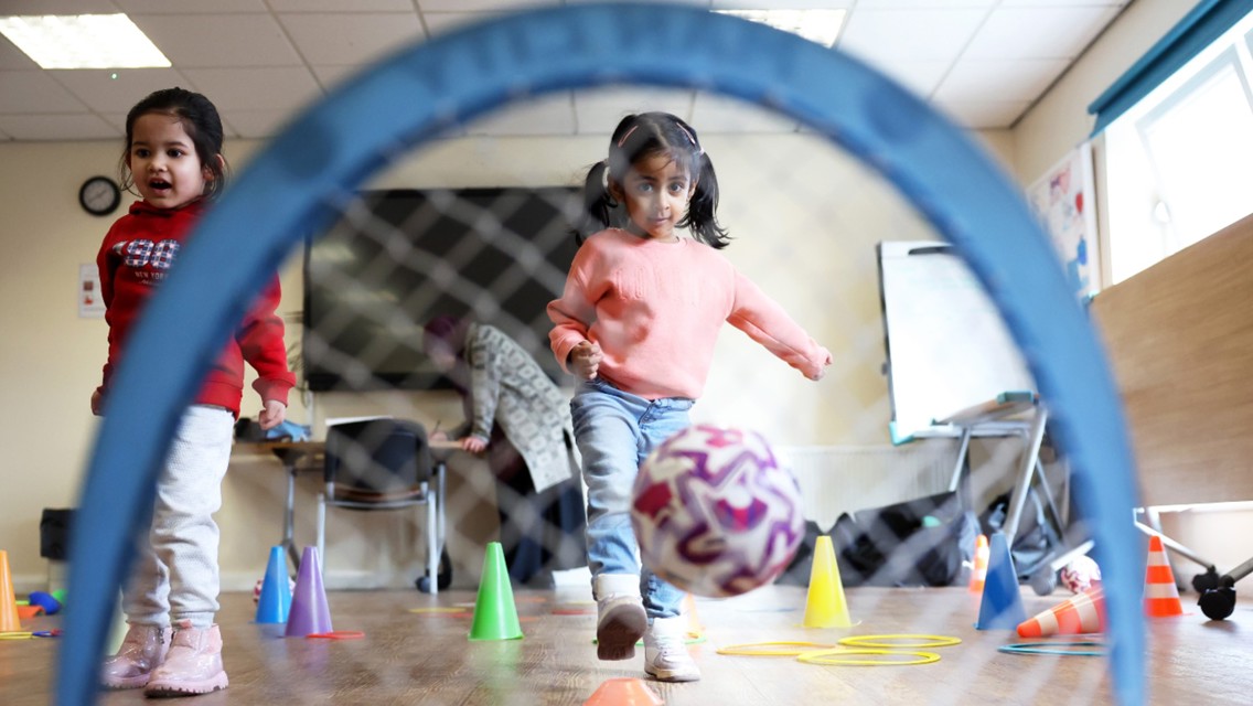Children participating in a play activity with a football inside a room with cones and rings.