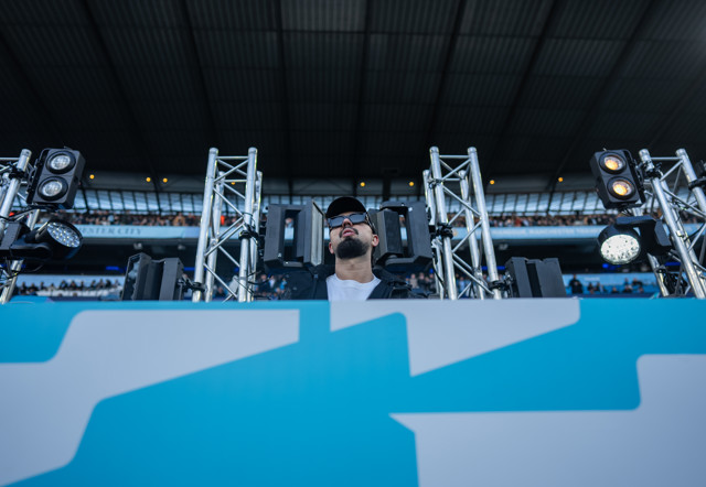 A blurred person is operating electronic equipment on a stage set up inside a stadium, lights and speakers are visible. The venue has an open roof.