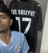 A person wearing a Manchester City jersey poses next to a hanging De Bruyne jersey with number 17.