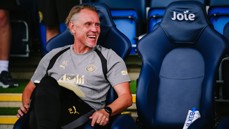 A man wearing Manchester City sports gear is sitting on a chair at a stadium, smiling and holding a notebook. The chair next to him has the word 'Joie' on it.
