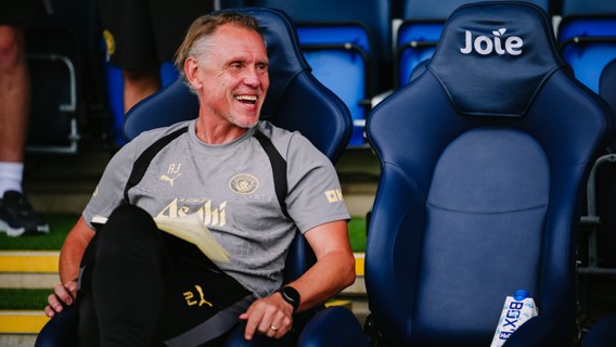 A man wearing Manchester City sports gear is sitting on a chair at a stadium, smiling and holding a notebook. The chair next to him has the word 'Joie' on it.