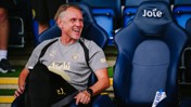 A man wearing Manchester City sports gear is sitting on a chair at a stadium, smiling and holding a notebook. The chair next to him has the word 'Joie' on it.