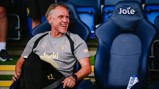 A man wearing Manchester City sports gear is sitting on a chair at a stadium, smiling and holding a notebook. The chair next to him has the word 'Joie' on it.