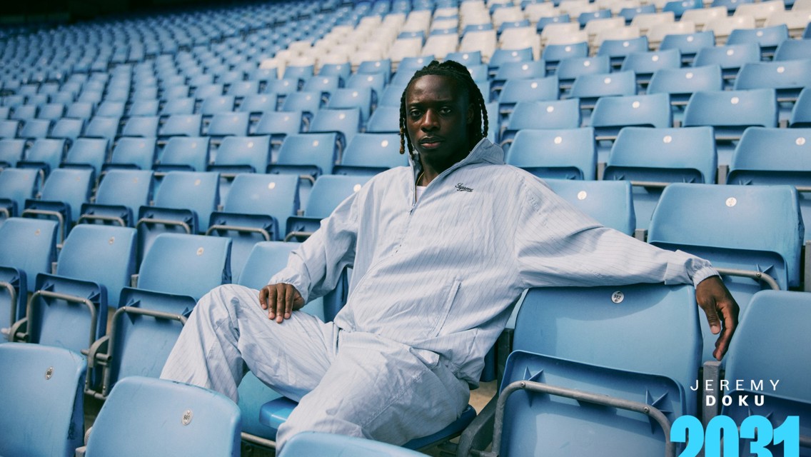 A man in a light blue tracksuit sits in a stadium with empty blue seats. A text overlay reads 'Jeremy Doku 2031' in the corner.