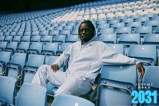 A man in a light blue tracksuit sits in a stadium with empty blue seats. A text overlay reads 'Jeremy Doku 2031' in the corner.